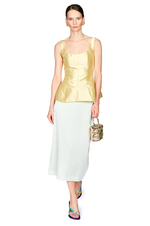 A model is shown from head to toe wearing a metallic yellow satin peplum top and a white midi skirt, holding a small patterned handbag against a plain white background.