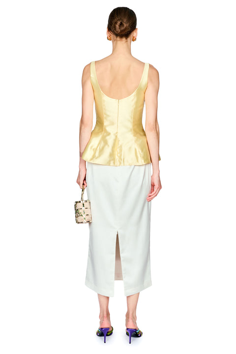 A model shown from the back wearing a yellow satin peplum top and a white midi skirt, holding a small patterned handbag against a white background.