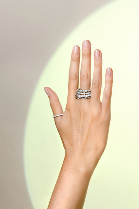 A woman's hand wearing a Spinelli Kilcollin ring that features a very thin platinum band with pave white diamonds, a very thick platinum band with pave white diamonds, a thin platinum band with pave white diamonds, and a thick platinum band with pave white diamonds, all connected by three platinum connector rings with pave white diamonds on her index finger. 
