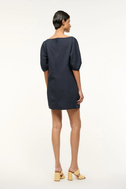 Back view of a woman wearing a navy, mini shift dress that features a boat neck and balloon half-sleeves, paired with tan heels. 