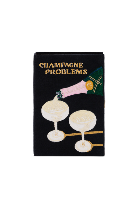 A black book clutch bag featuring a graphic illustration of a champagne bottle pouring into two champagne glasses, with the text 'CHAMPAGNE PROBLEMS' in gold lettering at the top.