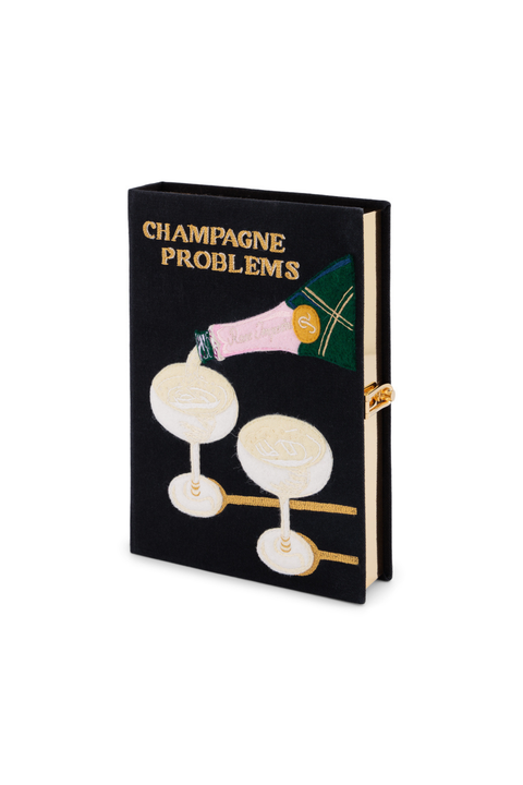 A black book clutch titled 'CHAMPAGNE PROBLEMS' with gold lettering and an illustration of champagne glasses and a pink champagne bottle, shown on a white background.