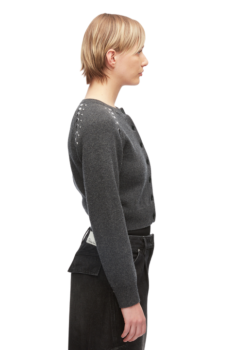 A side-profile view of a model wearing a dark gray knit cardigan with decorative silver stud embellishments along the shoulder yoke, paired with black jeans against a plain white background.