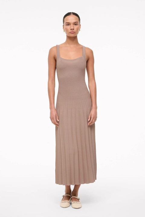 Woman wearing a sleeveless, taupe knit midi dress that features a scoop neck, fitted bodice, and pleated skirt, paired with cream flats. 