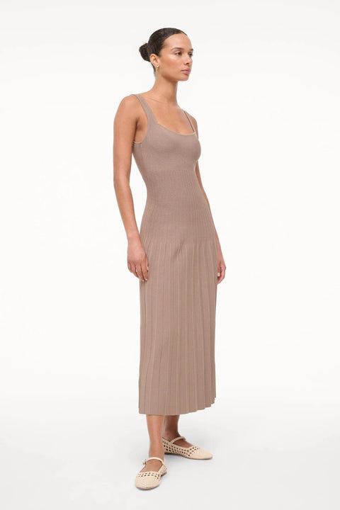 Side view of a woman wearing a sleeveless, taupe knit midi dress that features a scoop neck, fitted bodice, and pleated skirt, paired with cream ballet flats. 