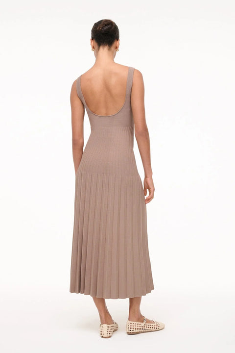 Back view of a woman wearing a sleeveless, taupe knit midi dress that features a low scoop back, fitted bodice, and pleated skirt, paired with cream ballet flats. 
