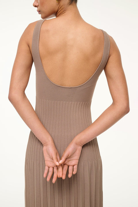 Close up of a woman wearing a sleeveless, taupe, knit midi dress that features  a low scoop back, ribbed, fitted bodice, and pleated skirt. 
