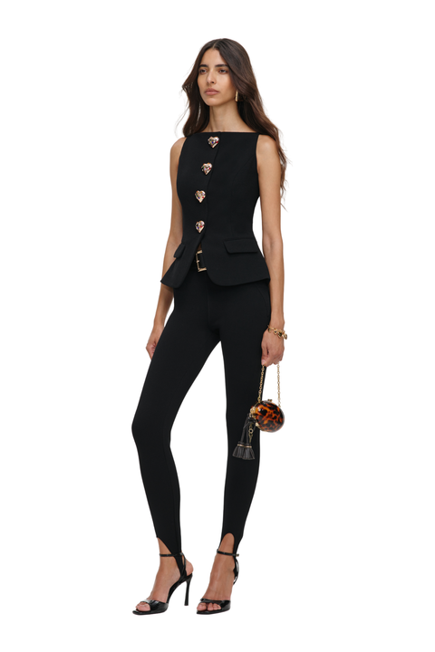 A woman modeling a black sleeveless tailored top and matching fitted pants, styled with black strappy heels and a small patterned handbag against a plain white studio background.