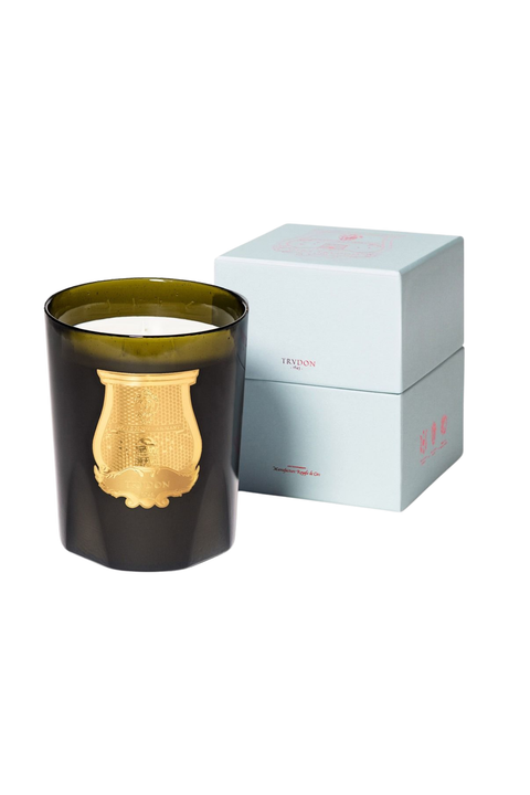 An amber green murano glass jar candle with a gold lyre-shaped gold emblem on the front, shown next to its light blue and pink branded box on a white background.