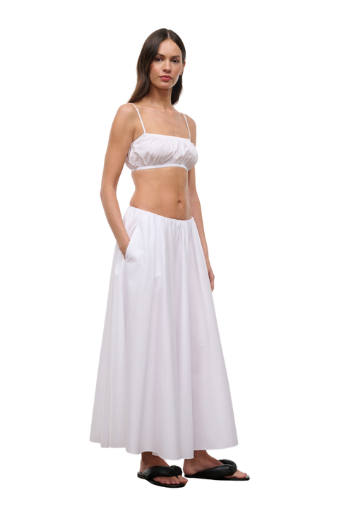 A woman modeling a white two-piece outfit consisting of a cropped camisole top with thin straps and a matching high-waisted maxi skirt with side pockets, paired with black slide sandals, on a solid white background.