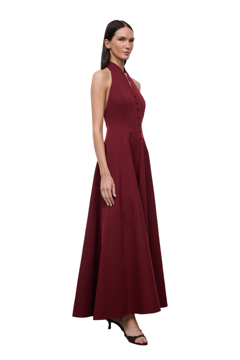 A woman modeling a sleeveless, high-neck, floor-length burgundy dress with a buttoned bodice, shown against a plain white background.