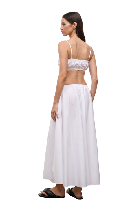 A woman shown from the back wearing a white two-piece set consisting of a ruffled crop top and a long pleated maxi skirt, paired with black slide sandals, against a plain white background.