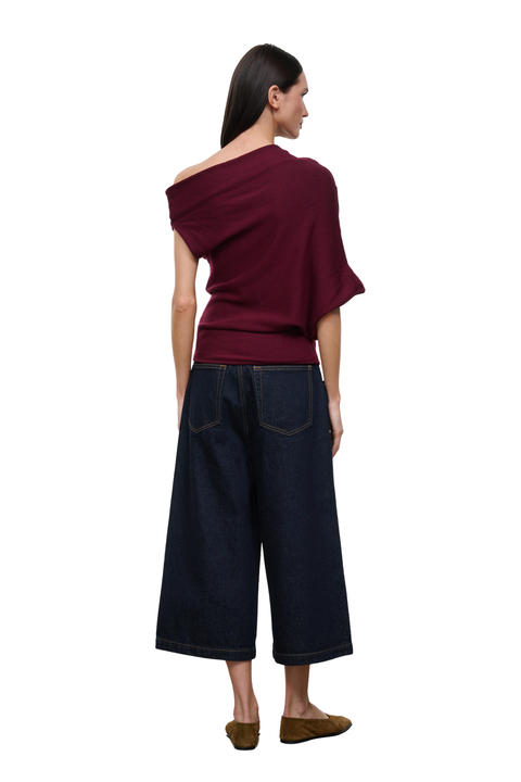 A woman shown from the back wearing a dark burgundy off-the-shoulder top and dark blue wide-leg cropped jeans, paired with brown slip-on shoes, against a plain white background.