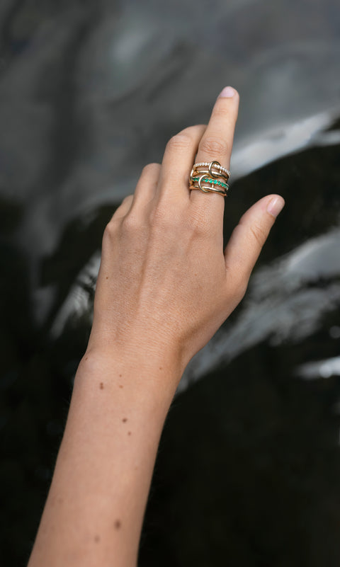 A woman's hand weawring a Spinelli Kilcollin ring that features a thin pave diamond yellow gold band, a think 18k yellow gold band, a thick yellow gold band pave with emeralds, and a thinner 18k yellow gold band, all connected with three 18k yellow gold connector rings on her index finger. 