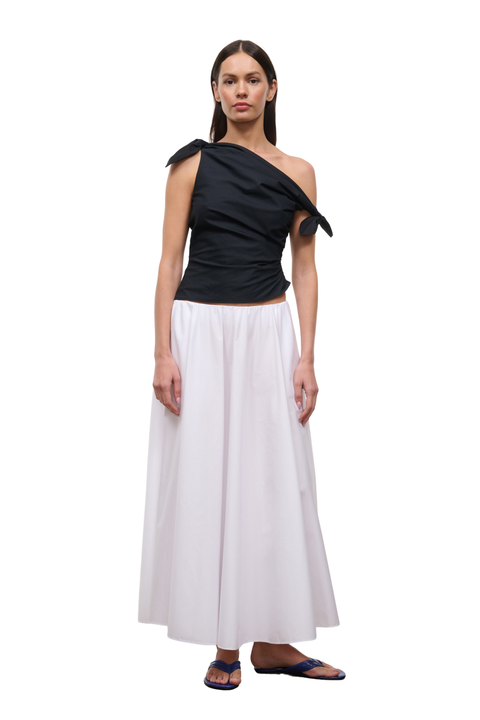 A woman modeling a two-piece outfit consisting of a black one-shoulder draped top with tie details at the shoulders and a long white skirt, paired with dark blue flip-flop sandals, against a plain white background.
