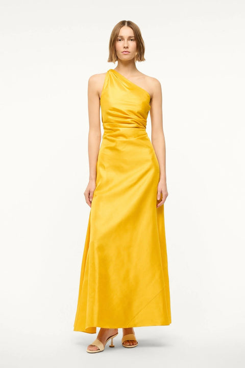 Woman wearing a yellow-gold silk maxi gown that features a one-shoulder detail with flattering draping at the bodice, paired with cream heels. 