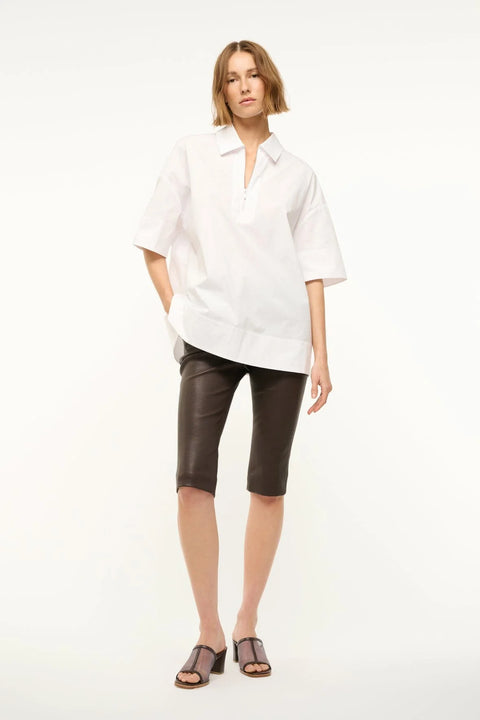Woman wearing a white oversized short sleeve, collared tunic that features a zipper detail at the neck, paired with black capris.  