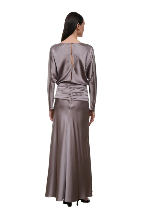 A woman viewed from the back wearing a long-sleeve satin-like maxi dress in a taupe-gray color, featuring a draped gathered waist and a back keyhole detail, set against a plain white background.