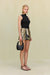 Side view of a woman wearing a black sleeveless top that features an asymmetrical high neck and ruched draping throughout the bodice, paired with an embellished mini skirt. 