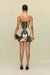 Back view of a woman wearing a strapless mini dress that features a black an tan portrait print depicting two faces looking at each other and holding a fruit in their hands, paired with a black clutch, black heels, and gold chunky jewelry. 