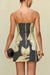 Up close back view of a woman wearing a strapless mini dress with a black and tan portrait pattern depicting two faces looking at each other and holding a fruit in their hands, paired with a black clutch and gold chunky jewelry. 