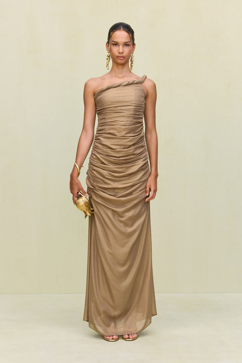 Woman wearing a gold metallic gown that features a hand rolled, one-shoulder neckline and intricate ruched draping throughout the body, paired with gold heels and accessories. 