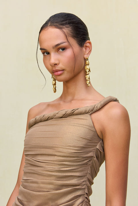 Close up of a woman wearing a gold metallic gown that features a hand rolled, one-shoulder neckline and ruched draping in the bodice, paired with gold statement earrings. 