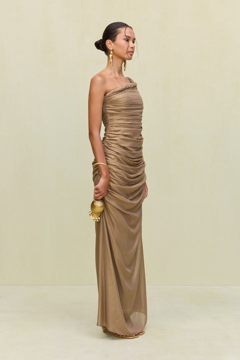 Side view of a woman wearing a gold metallic gown that features a hand rolled, one-shoulder neckline and intricate ruched draping throughout the body, paired with gold heels and accessories. 