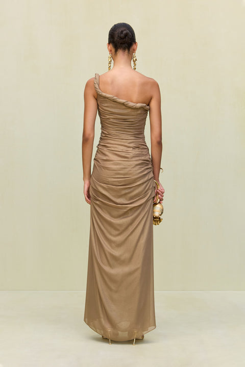 Back view of woman wearing a gold metallic gown that features a hand rolled, one-shoulder neckline and intricate ruched draping throughout the body, paired with gold heels and accessories. 
