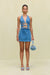 Woman wearing a blue denim mini dress that features a halter neck and torso cutouts embellished with gold buckle detailing, paired with a blue handbag and denim mules. 