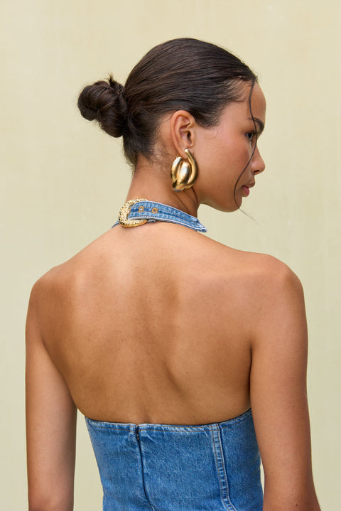 Up close back view of a woman wearing a blue denim mini dress that has a halter neck with an adjustable gold buckle detail at the back of the neck.