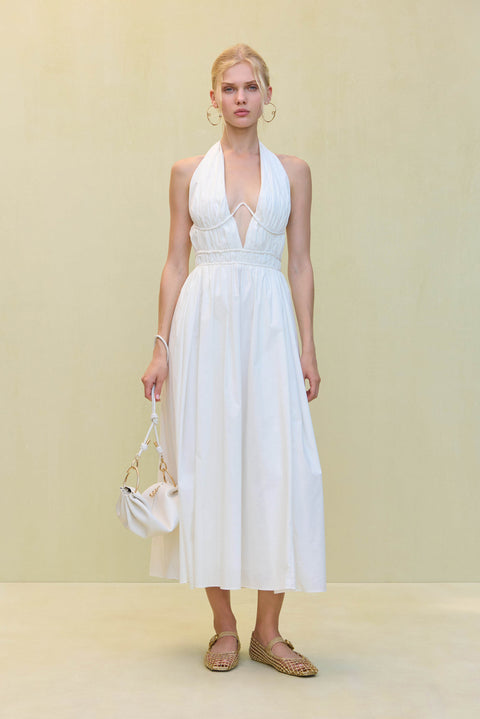 Woman wearing a white poplin midi dress that features a plunging halter neck and wire details at the bust, paired with a white handbag and tank sandals. 