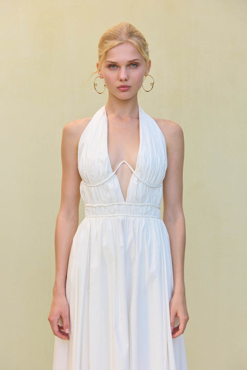 Close up of a woman wearing a white midi dress that features a plunging halter neck and wire details at the bust, paired with gold hoop earrings. 