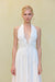 Close up of a woman wearing a white midi dress that features a plunging halter neck and wire details at the bust, paired with gold hoop earrings. 