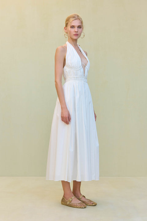 Side view of a woman wearing a white poplin midi dress that features a plunging halter neck and wire details at the bust, paired with gold flats. 