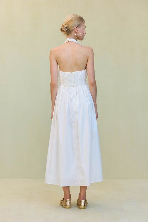 Back view of a woman wearing a white poplin midi dress that features a halter neck and open back, paired with old ballet flats. 