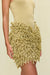 Detail shot of a woman wearing a gold knit mini skirt with a plunging waistline and all over gold knit looped fringe, paired with a gold bodysuit. 