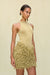 Close up side angle of a woman wearing a gold knit mini skirt with a plunging waistline and all over gold knit fringe, paired with a gold bodysuit and gold statement earrings. 