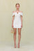 Woman wearing a white linen mini dress that features an off the shoulder neckline and gold hardware detailing at the bust.