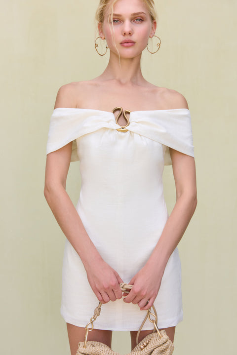 Close up of a woman wearing a white linen mini dress that features an off the shoulder neckline and gold hardware detailing at the bust.