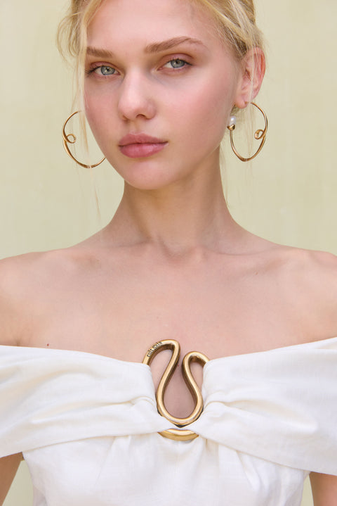 Detail shot of the off-the-shoulder neckline and abstract gold hardware at the bust of a white linen mini dress. 