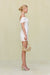 Side view of a woman wearing a white linen mini dress that features an off the shoulder neckline and gold hardware detailing at the bust.