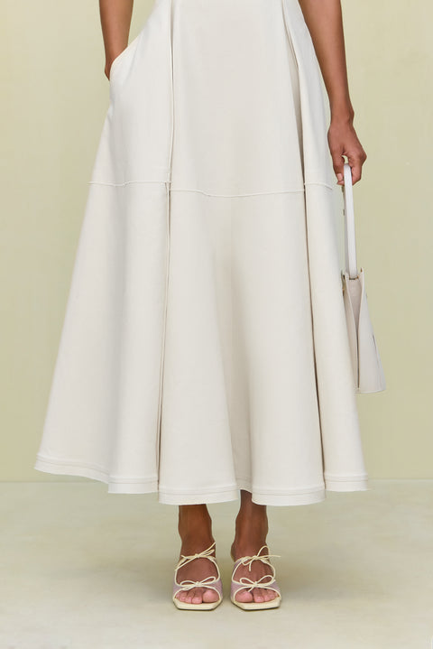 Close up of the full skirt of a white maxi dress that features pockets, paired with cream bow heels. 