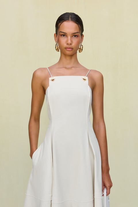 Close up of a woman wearing a white maxi dress that features a high square neck with gold-toned grommet details attaching the spaghetti straps, paired with chunky gold earrings. 