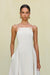 Close up of a woman wearing a white maxi dress that features a high square neck with gold-toned grommet details attaching the spaghetti straps, paired with chunky gold earrings. 