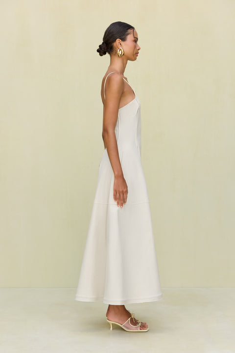 Side view of a woman wearing a white maxi dress that features a square neck with grommet strap details and a full skirt, paired with cream bow heels. 