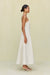 Side view of a woman wearing a white maxi dress that features a square neck with grommet strap details and a full skirt, paired with cream bow heels. 