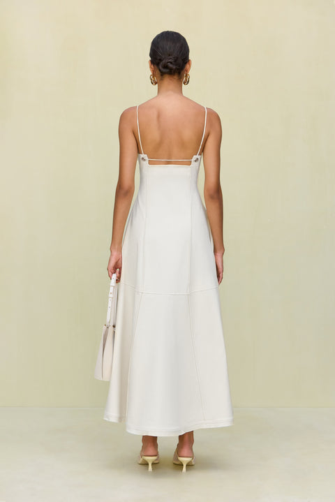 Back view of a woman wearing a white maxi dress that features a low square back with gold-toned grommet details attaching the spaghetti straps and a full skirt, paired with gold heels. 