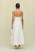 Back view of a woman wearing a white maxi dress that features a low square back with gold-toned grommet details attaching the spaghetti straps and a full skirt, paired with gold heels. 