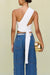 Back view of a woman wearing a white knit crop top that features an asymmetrical one shoulder detail that wraps into a draping strap. 